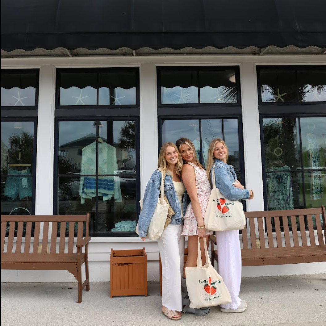 Three women standing outside a store named 'OOMA
 Boutique' holding bags.